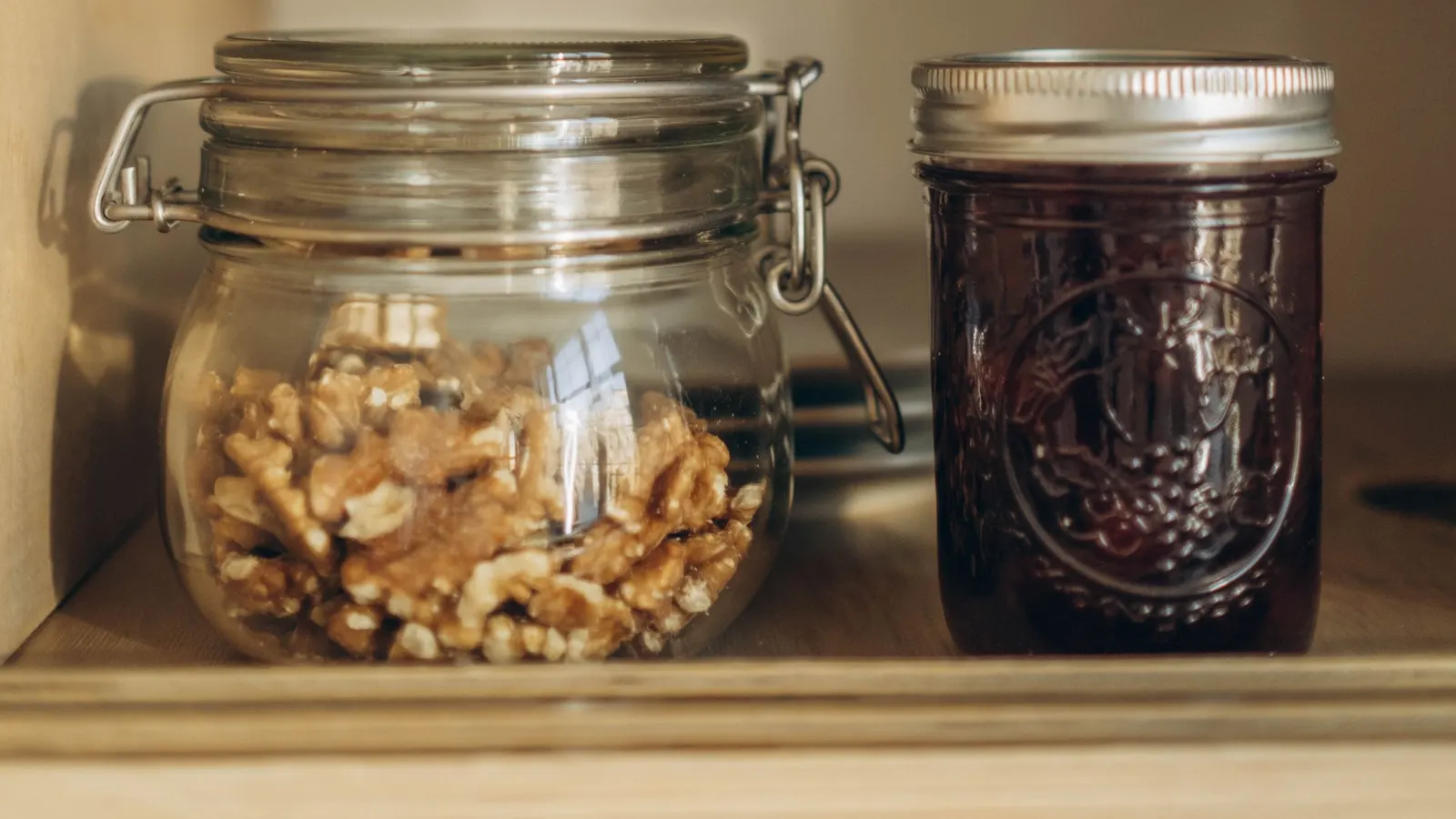 A glass jar of walnuts and a jar of preserves on a wooden pantry shelf
