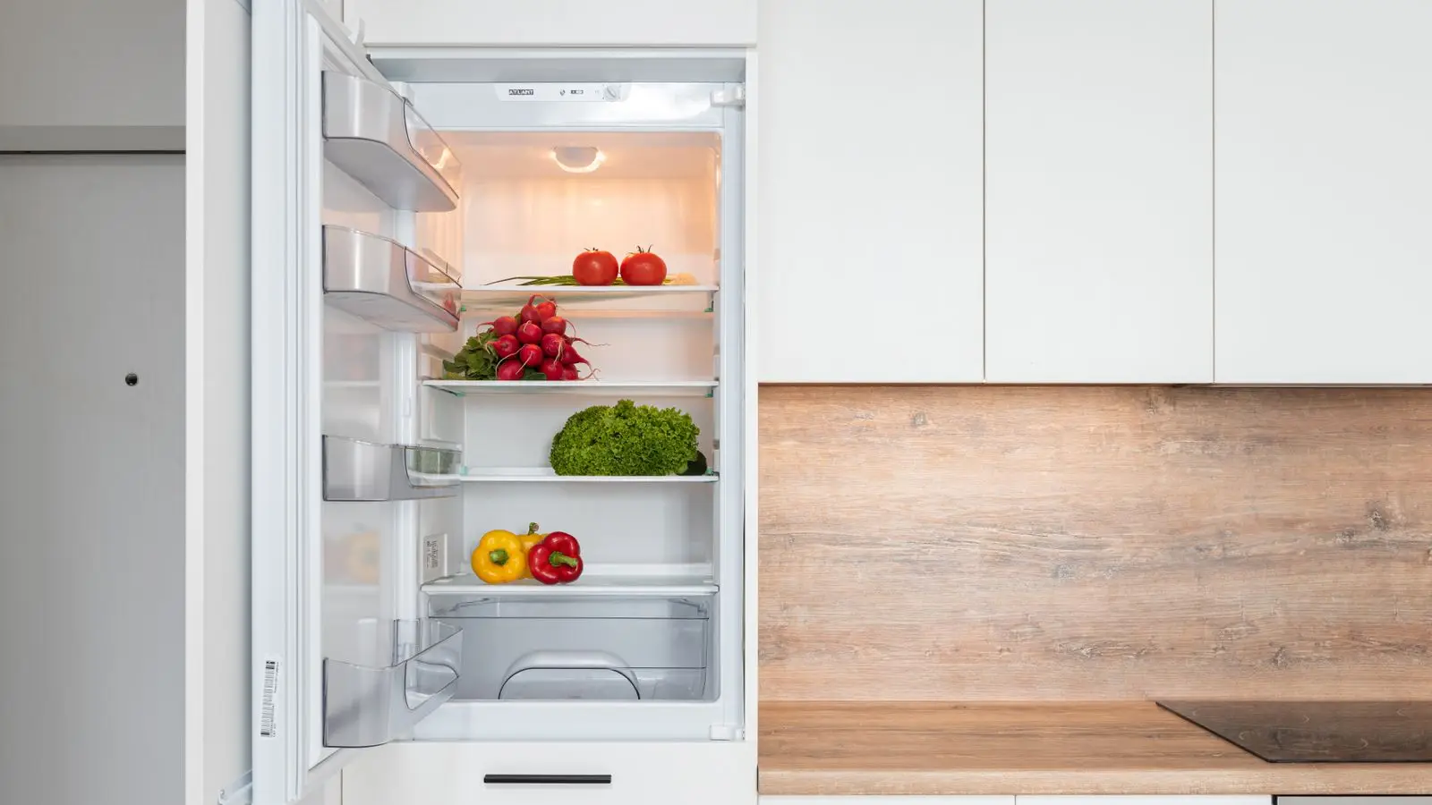 Open fridge in a white kitchen with tomatoes, radishes, lettuce and peppers on separate shelves