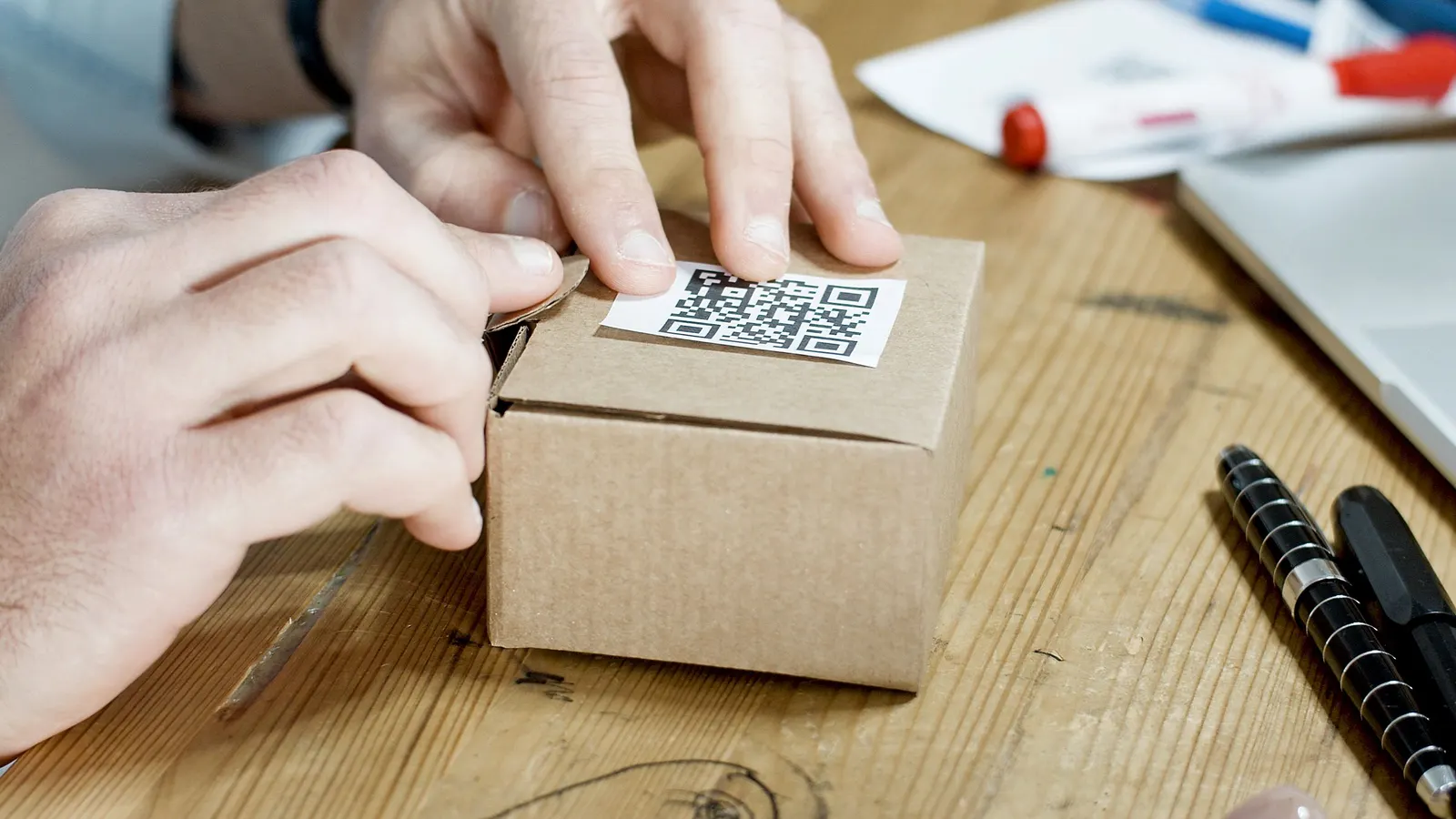 Hands pressing a printed QR code sticker onto a small cardboard box