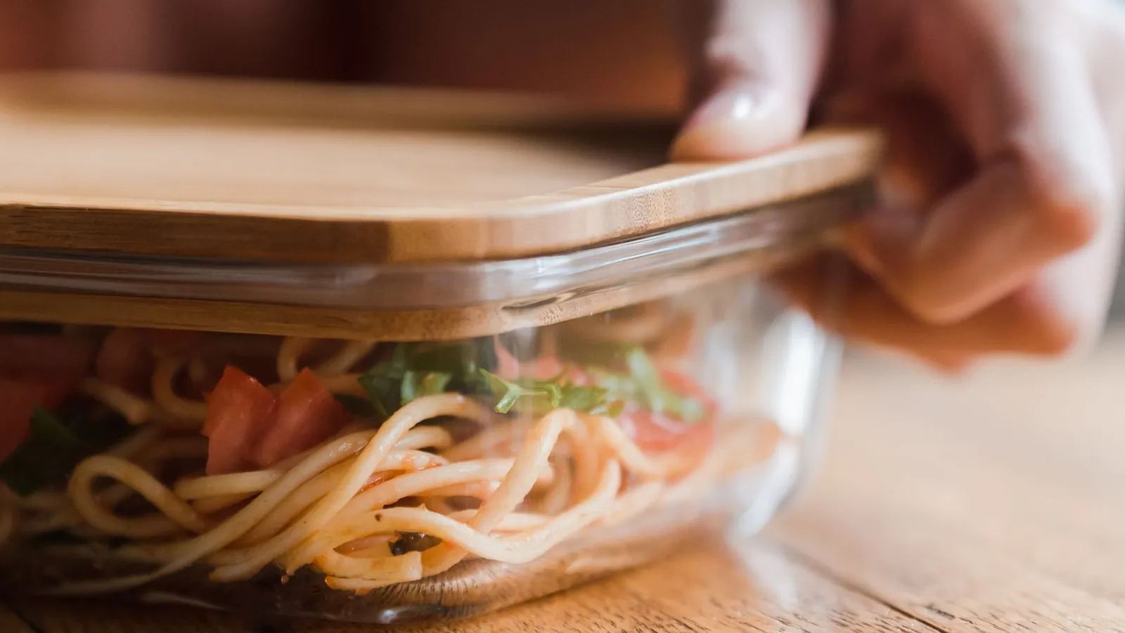 A hand pressing a wooden lid onto a glass container of leftover spaghetti with tomatoes and basil