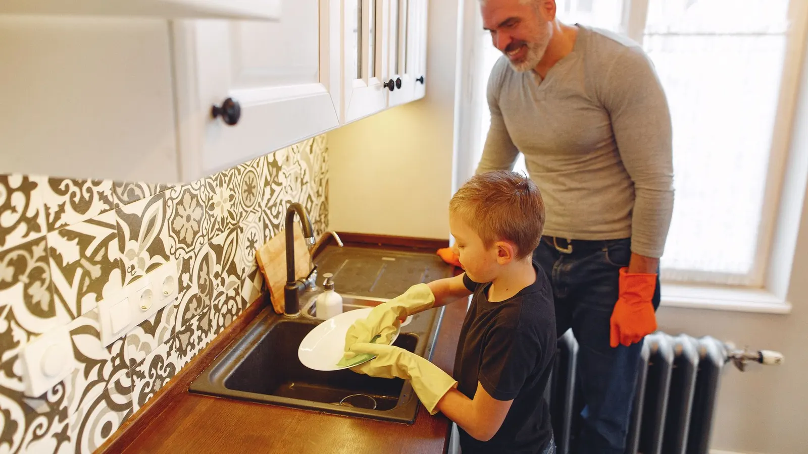 A boy in rubber gloves washes a plate at the kitchen sink while his smiling father watches